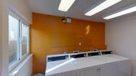 This single-floor laundry facility is designed for functionality and efficiency. The most striking visual element is the bold, warm orange accent wall, which adds a vibrant, energetic character to the otherwise utilitarian space. A row of modern, commercial-grade top-loading washing machines lines the room, each equipped with digital control panels, suggesting a contemporary and user-friendly setup. The machines are arranged in a neat, accessible row, maximizing the use of the available floor space.

Overhead, fluorescent lighting fixtures provide consistent, bright illumination throughout the room, ensuring excellent visibility during use. A security camera is mounted on the orange wall, indicating that the facility prioritizes the safety and security of its users.

The white ceiling and trim create a clean contrast against the orange wall, contributing to the space's crisp, well-maintained appearance. The layout is straightforward and practical, with clear access to each machine. Overall, the laundry facility presents a tidy, well-equipped environment that balances practicality with a distinct visual identity through its bold color choices.
