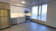 This contemporary single-floor space showcases a thoughtfully designed open-concept layout that seamlessly blends style and functionality. The kitchen is a standout feature, outfitted with a full suite of stainless steel appliances, including a range, over-the-range microwave, and dishwasher. Crisp white cabinetry extends from counter to ceiling, maximizing storage while contributing to a clean, modern aesthetic. A classic white subway tile backsplash adds a timeless touch, complementing the light-toned countertops and undermount sink. Light wood-tone flooring runs throughout the space, lending warmth and continuity to the overall design. Large floor-to-ceiling windows flood the interior with natural light and frame expansive views of the surrounding cityscape and beyond, creating a bright, airy ambiance. The open layout between the kitchen and living area promotes a fluid sense of space, making the interior feel generously proportioned. Recessed under-cabinet lighting enhances both the functionality and the atmosphere of the kitchen. Overall, the space strikes a balance between contemporary design and practical living, making it an inviting and well-appointed environment.