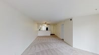 This single-floor residential unit offers a straightforward and functional layout designed for comfortable everyday living. The open-concept living area is finished with neutral-toned wall-to-wall carpeting that lends warmth and a cohesive feel throughout the space. Crisp white walls and a smooth ceiling contribute to a bright and airy ambiance, while a ceiling fan centrally positioned overhead provides both ventilation and a subtle decorative element.

The living area flows naturally toward the kitchen, which is defined by a raised peninsula counter — ideal as a casual breakfast bar or a convenient prep and serving surface. The kitchen features white cabinetry and appears to include standard appliances, maintaining the clean, uniform aesthetic of the unit. A sliding glass door adjacent to the kitchen allows natural light to filter into the space and suggests access to a patio or outdoor area.

A hallway off the main living area provides access to additional rooms, ensuring a logical separation between the living and private spaces. The unit's neutral color palette and practical design make it a versatile canvas suited to a variety of interior styles and furnishing preferences.