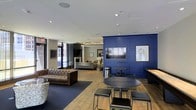This single-floor communal space is thoughtfully designed to blend functionality with a modern, upscale aesthetic. The layout is open and inviting, anchored by striking navy blue paneled accent walls that serve as a bold focal point throughout the room. A wall-mounted flat-screen television is prominently positioned against one of these navy walls, creating an ideal viewing area for residents or guests. The flooring transitions between warm hardwood and deep-toned carpet zones, helping to define distinct areas within the open plan. Seating options are varied and plentiful — from a tufted leather chesterfield sofa to compact round café-style tables paired with sleek stools, offering both casual lounging and informal gathering spaces. Recessed lighting throughout the ceiling provides even, ambient illumination, while large windows allow natural light to filter in, offering views of the surrounding urban streetscape. Toward the back of the space, an additional seating area with a fireplace and more relaxed furnishings creates a quieter retreat within the same room. Contemporary artwork adorns the walls, adding artistic character to the space. Overall, the design strikes a balance between professional polish and comfortable informality, making it a versatile and welcoming environment.