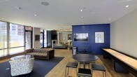 This single-floor communal space is thoughtfully designed to blend functionality with a modern, upscale aesthetic. The layout is open and inviting, anchored by striking navy blue paneled accent walls that serve as a bold focal point throughout the room. A wall-mounted flat-screen television is prominently positioned against one of these navy walls, creating an ideal viewing area for residents or guests. The flooring transitions between warm hardwood and deep-toned carpet zones, helping to define distinct areas within the open plan. Seating options are varied and plentiful — from a tufted leather chesterfield sofa to compact round café-style tables paired with sleek stools, offering both casual lounging and informal gathering spaces. Recessed lighting throughout the ceiling provides even, ambient illumination, while large windows allow natural light to filter in, offering views of the surrounding urban streetscape. Toward the back of the space, an additional seating area with a fireplace and more relaxed furnishings creates a quieter retreat within the same room. Contemporary artwork adorns the walls, adding artistic character to the space. Overall, the design strikes a balance between professional polish and comfortable informality, making it a versatile and welcoming environment.