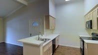 This single-story space is characterized by a clean, functional layout that connects the kitchen and entry areas in an open, airy arrangement. The kitchen serves as a central feature, anchored by a peninsula with a double-basin stainless steel sink and a pull-down faucet. Light-toned cabinetry lines both the upper wall-mounted units and the lower base cabinets, providing ample storage while maintaining a bright, neutral aesthetic. White countertops complement the cabinetry and extend across the peninsula, offering practical workspace. A dishwasher is integrated beneath the counter. The flooring throughout is a dark hardwood, creating a grounding contrast to the light walls and cabinetry. Crown molding runs along the upper walls, adding a refined architectural detail. The front entry is visible from the kitchen, featuring a white paneled door with an arched transom window that introduces natural light into the space. The walls are finished in neutral tones — a warm gray accent wall near the entry transitions to crisp white throughout the remainder of the space — contributing to an overall clean and move-in ready atmosphere.