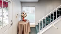 This single-floor commercial or residential common area serves as a functional and welcoming entry space. Upon entering through the paneled white door — framed with a multi-pane window that allows natural light to filter in — visitors are greeted by a warm and organized lobby environment. A round accent table draped in a neutral-toned cloth holds a decorative floral arrangement, adding a touch of charm near the entrance. A wall-mounted intercom or security panel is conveniently positioned beside the entry door.

The space features deep green carpeting throughout, extending up the carpeted staircase with white railings that leads to an upper level. A tall, lush decorative ficus tree in a woven basket planter stands prominently in the corner, adding a natural, welcoming element to the interior. Adjacent to the tree, a bulletin board displays various notices, certificates, and community information, suggesting a shared or managed property environment. Compliance postings are visible on the opposite wall, further indicating a professionally managed facility. A window at the staircase landing allows additional natural light to brighten the space. Overall, the area presents a tidy, organized, and inviting atmosphere suited for a multi-unit or office environment.