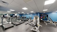 Located at 3214 Hewitt Ave in Silver Spring, this dedicated fitness center offers a well-organized and fully equipped exercise space designed to support a comprehensive workout routine. The single-floor layout is finished with durable rubber flooring throughout, providing both safety and comfort underfoot.

The cardio zone features a row of treadmills and elliptical trainers from True Fitness, all positioned along one side of the room for an efficient use of space. The strength training area is generously outfitted with a variety of Hoist and Life Fitness selectorized machines, including leg press, chest press, and cable systems, catering to users of all fitness levels. A dedicated free weights section includes a full rack of dumbbells and adjustable benches, while a wall-mounted rack holds medicine balls and stability balls for functional training.

Large wall-to-wall mirrors line multiple walls, visually expanding the space and allowing users to monitor their form during workouts. The room is illuminated by recessed panel lighting set within a drop-tile ceiling, creating a bright and even atmosphere. A wall-mounted television is also present, adding a convenient entertainment option. The space maintains a clean, professional aesthetic with bold blue accent walls contrasting against neutral white and grey tones throughout.