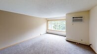 This single-floor residential unit offers a practical and comfortable layout across several distinct living areas. The living room is a spacious, carpet-floored space bathed in natural light through a large picture window with warm wood trim, complemented by baseboard heating and a wall-mounted air conditioning unit for year-round comfort. The textured ceilings are consistent throughout, adding a classic residential character to the space.

The kitchen is well-equipped and efficiently designed, featuring white cabinetry with warm oak accents, a double-basin sink, a dishwasher, refrigerator, and electric range. Light tile flooring and bright overhead fluorescent lighting make the kitchen feel clean and functional. A peninsula counter provides additional workspace and casual dining potential.

The bedrooms are carpeted and bright, each featuring wood-trimmed windows that frame views of mature, leafy trees outdoors. One bedroom includes an open walk-in closet with built-in shelving and hanging rods, offering ample storage. The hallway area features tile flooring and a large double-door closet for additional storage.

The bathroom includes a soaking tub with a towel bar, a long vanity with oak cabinetry and a wide mirror, and tile flooring, creating a clean and practical space. Overall, the unit presents a cohesive, functional interior with consistent finishes throughout.