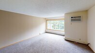 This single-floor residential unit offers a practical and comfortable layout across several distinct living areas. The living room is a spacious, carpet-floored space bathed in natural light through a large picture window with warm wood trim, complemented by baseboard heating and a wall-mounted air conditioning unit for year-round comfort. The textured ceilings are consistent throughout, adding a classic residential character to the space.

The kitchen is well-equipped and efficiently designed, featuring white cabinetry with warm oak accents, a double-basin sink, a dishwasher, refrigerator, and electric range. Light tile flooring and bright overhead fluorescent lighting make the kitchen feel clean and functional. A peninsula counter provides additional workspace and casual dining potential.

The bedrooms are carpeted and bright, each featuring wood-trimmed windows that frame views of mature, leafy trees outdoors. One bedroom includes an open walk-in closet with built-in shelving and hanging rods, offering ample storage. The hallway area features tile flooring and a large double-door closet for additional storage.

The bathroom includes a soaking tub with a towel bar, a long vanity with oak cabinetry and a wide mirror, and tile flooring, creating a clean and practical space. Overall, the unit presents a cohesive, functional interior with consistent finishes throughout.