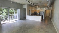This single-floor space showcases an industrial-modern aesthetic defined by its polished concrete floors and exposed concrete ceiling, which together lend a raw, urban character to the interior. The open-concept layout seamlessly connects the living area to the kitchen, creating a spacious, unobstructed flow throughout. A sliding glass door along one wall floods the living area with natural light and provides direct access to an outdoor deck or balcony, where views of surrounding greenery are visible.

The kitchen serves as a central feature, anchored by a peninsula with a dark countertop that offers both workspace and a natural dividing point between cooking and living areas. Light wood cabinetry lines the kitchen walls, complemented by black appliances including a range and a stainless steel refrigerator. A track lighting system mounted to the exposed ceiling runs across the space, providing adjustable, functional illumination, while pendant lights above the kitchen add a refined touch. The combination of the raw concrete surfaces with the warm wood cabinetry and modern fixtures creates a balanced, loft-inspired ambiance throughout the space.