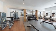 This health club is a spacious, single-level fitness facility thoughtfully designed to accommodate a comprehensive range of workout styles. The interior features warm hardwood flooring throughout, crisp white walls, and recessed lighting complemented by ceiling fans, creating a clean, well-ventilated environment conducive to exercise.

The facility is organized into distinct training zones. The cardio area hosts a row of treadmills lined along a mirrored wall with a mounted flat-screen television, flanked by windows fitted with blinds that allow natural light while maintaining privacy. Adjacent to this, elliptical trainers and additional cardio machines are positioned near windows offering outdoor views.

The strength training section is anchored by wood-accented structural columns and features a well-stocked dumbbell rack, multiple cable and weight machines, a Smith machine, and dedicated benching stations set atop rubber-padded flooring. Mirrors throughout amplify the sense of space and support proper form.

A functional training area near large glass double doors — which open to an outdoor terrace — includes plyo boxes, stability balls, medicine balls, and resistance equipment. A motivational wall graphic reading "Don't Wish For It, Work For It" adds an energetic focal point near the entrance. The facility's open layout and multiple zones make it suitable for a wide variety of fitness routines.