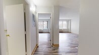 Located in Hartford, CT, this single-floor unit offers a clean, functional layout with a bright and neutral aesthetic throughout. The living and bedroom areas are finished with multi-toned hardwood-style plank flooring that adds warmth and visual character to each room. Large windows fitted with vertical blinds allow generous natural light to fill the space while providing views of the surrounding urban streetscape, and wall-mounted HVAC units ensure year-round comfort. The open living area connects seamlessly to a pass-through kitchen, which features white cabinetry, gray laminate countertops, and a full suite of white appliances including a refrigerator, electric range, built-in microwave, and dishwasher. The kitchen is finished with tile flooring, providing a practical and easy-to-maintain surface. The bedroom is well-proportioned and includes a built-in closet with double bifold doors and shelving, offering ample storage. A dedicated hallway leads to a full bathroom appointed with white tile flooring, a soaking tub, a single vanity with a mirrored cabinet, and a toilet. The unit also features a utility panel accessible from the entry corridor. The overall design is clean and move-in ready, well-suited for urban living in the heart of Hartford.