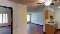 This single-floor space offers a straightforward and functional layout designed for comfortable everyday living. The main living area is highlighted by rich, dark hardwood flooring that runs throughout, providing a warm and grounded aesthetic. A ceiling fan is centrally positioned overhead, complementing the textured ceiling that adds subtle dimension to the space. Natural light filters in through a large window dressed with vertical blinds, brightening the open area and creating an inviting atmosphere. The neutral white walls offer a clean, versatile backdrop that easily accommodates a variety of furnishings and décor styles. A partial wall divides the living space from the adjacent kitchen area, where warm-toned wooden cabinetry is visible, hinting at a practical and efficiently organized kitchen. The dark baseboard trim provides a defined contrast against the light walls, lending a polished finishing touch to the interior. Overall, the space presents a simple, well-maintained environment with classic residential features that balance functionality and comfort in a compact, single-level footprint.
