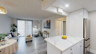 This efficiently designed 1-bedroom unit offers a thoughtfully organized layout across a single floor, encompassing a combined dining room, kitchen, and living area, one bedroom, a hallway, and a closet. The open-concept kitchen is a standout feature, outfitted with sleek white high-gloss cabinetry, stainless steel appliances, a subway tile backsplash, and a striking quartz-topped island with seating — ideal for casual dining or meal prep. The kitchen flows seamlessly into the dining nook, which features warm lighting and a dedicated closet, and continues into the living room. The living area is bright and airy, benefiting from large sliding glass doors dressed with vertical blinds that allow abundant natural light and provide access to an outdoor balcony with exterior views. Light grey wood-look flooring runs throughout, giving the space a clean, cohesive aesthetic. The bedroom is a comfortable and private retreat, featuring a generously sized window with outdoor views, vertical blinds, and the same consistent flooring. The hallway connects all areas smoothly, with a welcoming entryway accented by wall art and a console table. Overall, the unit presents a modern, neutral palette with clean lines and practical design.