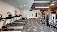 This spacious fitness center is designed to support a comprehensive workout experience across multiple training zones. The interior is defined by bold red columns and accent walls that contrast with neutral grey tones, creating an energetic and motivating atmosphere. Patterned carpet tile in grey and white covers the floor throughout, adding texture and visual interest underfoot.

The cardio zone is lined with a row of TRUE-brand treadmills and stationary bikes positioned along a wall featuring a large painted silhouette of a runner, reinforcing the space's athletic identity. Recessed lighting illuminates the area evenly, while windows and glass double doors along one wall invite natural light and provide outdoor views.

The strength training area is anchored by bold red columns and includes a full dumbbell rack, kettlebells, a cable machine, a multi-station weight machine, TRX suspension trainers, and a stability ball. Large wall-mounted mirrors expand the sense of space and allow users to monitor their form. A mounted flat-screen television serves the strength zone as well.

The coffered ceiling with dark-toned beams adds architectural dimension, and the overall layout ensures ample open floor space for functional and free-form training.