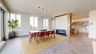Located at 1170 Hancock St in Quincy, MA, this thoughtfully designed single-floor space blends contemporary aesthetics with functional living. Throughout, striking chevron-patterned hardwood flooring ties the open-plan layout together, creating a cohesive and polished look.

The dining area is a standout, anchored by a sleek linear fireplace framed by a floor-to-ceiling decorative tile surround set within warm wood paneling. Sculptural circular pendant lights hang above a wooden dining table surrounded by terracotta-hued chairs, all set atop a woven yellow area rug. Expansive windows flood the space with natural light and offer open views of the surrounding Quincy skyline.

The kitchen is equally impressive, featuring light wood cabinetry, a textured stone backsplash, open display shelving, and a generous white-topped island with bar seating. Globe pendant lights and a window overlooking the exterior further brighten the space.

The living room offers a relaxed atmosphere with a modular sofa, a colorful large-scale art piece, and globe ceiling fixtures. Frosted glass partitions add a layer of architectural interest while maintaining visual connectivity throughout the unit.

A sliding glass door in the living area opens to a private outdoor terrace, extending the space and providing sweeping open-air views.
