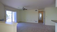 This single-floor residence offers a clean, functional layout designed for comfortable everyday living. The main living area is generously proportioned, featuring wall-to-wall carpeting that adds warmth and a soft, neutral aesthetic throughout. A ceiling fan is centrally positioned overhead, providing both air circulation and a practical touch to the space. Soft yellow-toned walls create a bright, welcoming atmosphere that pairs well with the natural light filtering in through the sliding glass door along one wall, which also provides access to an outdoor area. Ventilation is well accounted for, with multiple floor-level and ceiling-mounted HVAC vents visible throughout the room. A structural column detail near the entry hints at an open-concept transition into an adjacent area, such as a kitchen or dining space. A hallway leading toward what appears to be a front entry door with a window above it connects the living area to the rest of the home, suggesting a thoughtfully organized floor plan. The overall ambiance is light, airy, and move-in ready, with a neutral palette that offers flexibility for a variety of interior design styles.