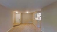 This single-floor space offers a clean, open layout designed for comfortable everyday living. The main living area is finished with light-toned tile flooring that extends throughout, creating a sense of continuity and spaciousness. Neutral beige walls are complemented by crisp white trim and baseboards, lending the space a bright and welcoming ambiance.

A defining architectural detail is the kitchen pass-through window, which opens from the living area into the kitchen, promoting an connected, airy feel between the two spaces. The kitchen, visible through the pass-through, features white cabinetry and appears well-appointed for functional use.

Overhead, flush-mount ceiling light fixtures provide consistent illumination across the living area, ensuring a well-lit environment. A white paneled door leads to an adjacent room or closet, adding to the practical layout of the space.

The overall design embraces a simple, understated aesthetic with its warm neutral palette, clean lines, and uncluttered floor plan. The combination of light flooring, soft wall tones, and thoughtful layout details make this a versatile and inviting space suited for a variety of uses.