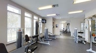 This fitness center offers a dedicated space for comprehensive workouts in a clean and modern environment. The main exercise area is outfitted with a multi-function strength training machine, providing users with a variety of resistance training options. A wall-mounted flat-screen television adds an entertainment element to workouts. The space is brightened by multiple windows fitted with blinds, allowing abundant natural light to filter in while maintaining privacy. Overhead fluorescent lighting ensures the area remains well-lit throughout the day. The flooring features durable, rubberized gym-grade material, ideal for heavy equipment and high-impact activity. A large wall mirror extends the visual depth of the space and allows users to monitor their form during exercise. Beyond the main area, an adjoining room is visible, housing additional cardio equipment such as stationary bikes and treadmills, effectively expanding the overall workout capacity of the facility. The neutral, light-toned walls and clean layout contribute to an open and focused atmosphere. Ventilation is provided through ceiling-mounted HVAC vents, ensuring a comfortable environment during physical activity. Overall, this fitness center is thoughtfully arranged to support a full range of exercise routines.
