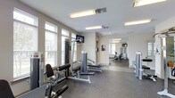 This fitness center offers a dedicated space for comprehensive workouts in a clean and modern environment. The main exercise area is outfitted with a multi-function strength training machine, providing users with a variety of resistance training options. A wall-mounted flat-screen television adds an entertainment element to workouts. The space is brightened by multiple windows fitted with blinds, allowing abundant natural light to filter in while maintaining privacy. Overhead fluorescent lighting ensures the area remains well-lit throughout the day. The flooring features durable, rubberized gym-grade material, ideal for heavy equipment and high-impact activity. A large wall mirror extends the visual depth of the space and allows users to monitor their form during exercise. Beyond the main area, an adjoining room is visible, housing additional cardio equipment such as stationary bikes and treadmills, effectively expanding the overall workout capacity of the facility. The neutral, light-toned walls and clean layout contribute to an open and focused atmosphere. Ventilation is provided through ceiling-mounted HVAC vents, ensuring a comfortable environment during physical activity. Overall, this fitness center is thoughtfully arranged to support a full range of exercise routines.