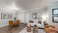 This single-floor residence offers a thoughtfully designed layout that balances comfort and style. The living area serves as the heart of the home, featuring dark hardwood-style flooring, crown molding, and a ceiling fan. Natural light fills the space through multiple windows dressed with slatted blinds, and a glass-paned exterior door adds additional brightness. The living room comfortably accommodates a seating arrangement anchored by a patterned area rug, with ample wall space for artwork.

Adjacent to the living area, the dining space features a round table with seating for multiple guests and convenient access to double-door storage. The kitchen is equipped with light wood cabinetry, black appliances including a range and refrigerator, and granite countertops that extend to a peninsula, providing additional prep and serving space.

The bedroom is carpeted for warmth and comfort, featuring a ceiling fan, a window with natural views, and generous closet access. The adjoining bathroom showcases rich dark wood vanity cabinetry topped with granite countertops, offering practical storage and a polished finish.

Throughout the residence, soft gray walls and consistent finishes create a cohesive, modern aesthetic, making this a comfortable and well-organized living space.