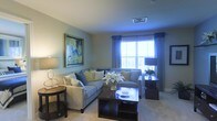 This single-floor space offers a thoughtfully designed interior that balances comfort and style. The living room serves as the centerpiece, showcasing a generous layout with a plush sectional sofa arranged around a dark-stained wood coffee table, creating an inviting area for relaxation and gathering. Soft, neutral-toned walls complement the light carpeted flooring, lending the space a warm and cohesive aesthetic. A large framed abstract artwork adds a vibrant focal point to the room, while carefully placed table lamps provide warm ambient lighting throughout. The space benefits from abundant natural light streaming through large windows dressed with elegant blue drape curtains, offering views of the surrounding greenery outside. The ceiling features a standard HVAC vent, ensuring year-round comfort. The overall ambiance is polished yet welcoming, with decorative touches such as a floral centerpiece on the coffee table and coordinated accent pillows that add layers of texture and color. The single-floor layout promotes easy flow and accessibility, making the space both functional and visually appealing for everyday living.