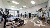 Wellington Place Performance is a comprehensive 1,800-square-foot fitness facility spanning a single floor and divided into distinct training zones designed to support a wide range of athletic and wellness goals. The main cardio and strength area features patterned geometric carpeting and a dropped ceiling with recessed panel lighting, creating a bright and well-lit environment. This zone is lined with large wall mirrors that visually expand the space, and is equipped with a full array of Life Fitness cardio machines — including treadmills, ellipticals, stair climbers, and rowing machines — alongside a robust selection of Life Fitness selectorized strength machines for targeted muscle training. A separate functional training zone features artificial turf flooring and white walls, giving it a clean, athletic aesthetic. This area is outfitted with a Hammer Strength power rack, an adjustable bench on a hardwood lifting platform, kettlebells, resistance bands, a heavy bag, cable machines, a dumbbell rack, and weighted sleds — accommodating strength, power, and conditioning workouts. The facility's bold branded mural prominently displays the Wellington Place Performance name, reinforcing its identity as a dedicated performance training space. Together, the zones provide a complete, well-equipped environment for users of all fitness levels.