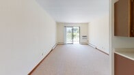 This single-floor unit offers a practical and straightforward layout across several distinct areas. The living room is a generously sized, open space finished with neutral beige carpeting and cream-colored walls trimmed with rich dark wood baseboards. A sliding glass door floods the room with natural light and provides direct access to an outdoor area, offering views of surrounding greenery. A wall-mounted air conditioning unit and baseboard heating ensure year-round comfort.

A pendant light fixture marks the transition to a secondary room or dining area, which shares the same neutral carpet and warm trim. This space connects conveniently to the kitchen, which features warm-toned wood cabinetry, a coil-burner electric range with an overhead ventilation hood, a stainless steel sink, and light laminate countertops — providing all the essentials in a compact, efficient arrangement.

The bedroom is a well-proportioned room with the same beige carpeting, cream walls, and dark baseboards. A wide horizontal window with blinds allows natural light while maintaining privacy, and baseboard heating adds to the room's comfort.

The bathroom is accessed through a wood-framed door and features small-format mosaic tile flooring, a tub and shower combo enclosed by sliding glass doors, and a standard toilet — completing this functional and cohesive unit.