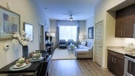 This single-floor residence offers a thoughtfully designed open-concept layout that seamlessly connects the living and dining areas into one cohesive, functional space. The living area serves as the centerpiece of the home, anchored by a large window dressed with elegant floor-to-ceiling drapes that flood the room with natural light. A ceiling fan overhead provides both practicality and a subtle design touch. The space is furnished with a comfortable sectional sofa, a glass-top coffee table, and a pair of accent chairs, creating a balanced and inviting seating arrangement ideal for both relaxing and entertaining. Hardwood-style flooring runs throughout, lending warmth and a clean, contemporary aesthetic to the interior. Neutral gray walls provide a versatile backdrop that complements the carefully curated décor, including wall art and decorative accents that add personality without overwhelming the space. Adjacent to the living area, the dining space accommodates a dark-toned table with upholstered seating, offering a defined area for meals. A paneled interior door adds an architectural detail that enhances the overall refined character of the space. Together, the design elements create a bright, modern, and welcoming atmosphere throughout.