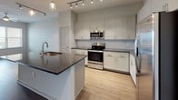 This single-story, one-bedroom, one-bathroom residence is thoughtfully designed with contemporary finishes and an efficient, open layout. The kitchen serves as a central highlight, featuring crisp white shaker-style cabinetry, dark granite countertops, a large center island with an undermount sink, stainless steel appliances including a refrigerator with ice maker, electric range, and microwave, plus a tile backsplash and track lighting overhead. Pendant lights above the island add a stylish touch. Light wood-style flooring runs through the kitchen area, transitioning to darker hardwood-style flooring in the living area, which is brightened by windows with outdoor views and a ceiling fan. The bedroom offers plush carpeting, a large window allowing ample natural light, and a ceiling fan for comfort. The bathroom is well-equipped with a granite-topped vanity featuring gray cabinetry, a large framed mirror, and a full soaking tub with a shower. Dark hardwood-style flooring continues through the bathroom. An in-unit laundry area is also visible, adding everyday convenience to this modern, move-in-ready residence.