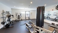 This fitness room offers a dedicated space for strength and resistance training, thoughtfully designed with functionality in mind. The room features clean white walls, recessed ceiling lighting, and durable rubber flooring that provides both safety and comfort during workouts. A variety of equipment is arranged throughout the space, including a cable crossover machine, a weight bench with rack, a leg press or seated resistance machine, and additional benches for varied exercises. A wall-mounted workout reference chart assists users in performing exercises correctly. Natural light filters in through a window along one wall, contributing to a bright and energizing atmosphere. A mirror is positioned along one side of the room, a practical feature for monitoring form during exercise. The layout allows for adequate spacing between equipment, enabling users to move freely and safely. The overall design is utilitarian yet polished, creating an environment well-suited for focused, effective training sessions.