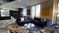 This elegantly designed single-floor space exudes a contemporary sophistication that is immediately apparent upon entry. The centerpiece of the space is a luxurious lounge area anchored by a deep navy sectional sofa adorned with an eclectic mix of accent pillows in gold, plum, and patterned tones, creating a rich and inviting atmosphere. A sleek glass and chrome coffee table sits atop a geometric-patterned area rug, adding a polished, modern touch to the seating arrangement. A tufted leather ottoman in warm cognac provides additional seating and visual contrast. Large floor-to-ceiling windows line the walls, flooding the interior with generous natural light and offering lush, verdant views of mature trees and landscaping beyond. The dark feature wall housing a flat-screen television provides a dramatic focal point, complemented by an elegant chandelier that adds a refined, upscale quality to the room. Curated wall art in warm amber and earthy tones punctuates the neutral palette, while recessed ceiling lighting enhances the overall ambiance. The thoughtful layering of textures, materials, and lighting throughout this space creates a cohesive and welcoming environment that balances comfort with contemporary design.