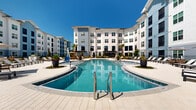 Located at 34 Ramsey Branch Rd in Freeport, Florida, this outdoor amenity space is a well-appointed residential community hub designed for relaxation and recreation under the Florida sun. The centerpiece is a generously sized, freeform swimming pool with crystal-clear blue water, multiple entry points including stainless steel handrails, and a shallow wading area. The pool is framed by an expansive, light-toned paved deck furnished with rows of chaise lounges, shade umbrellas, and seating areas ideal for sunbathing and socializing.

The surrounding landscape is thoughtfully designed with tropical plantings, including palm trees, ornamental grasses, flowering shrubs, and mulched garden beds that add texture and a lush, resort-like atmosphere. Raised planter islands within the pool deck area serve as decorative focal points. A dedicated outdoor grilling and dining station is integrated into the amenity space, providing a convenient area for outdoor cooking and entertaining.

The community is enclosed by decorative black iron fencing, with manicured lawns extending beyond the pool courtyard. Multi-story residential buildings with a contemporary coastal aesthetic — featuring white and blue-grey horizontal siding, large windows, and private balconies — surround the amenity area, creating an intimate and sheltered outdoor environment open to expansive Florida skies.