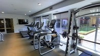 This fitness center is designed to provide a comprehensive workout environment within a single-level layout. The space is outfitted with a robust selection of exercise equipment, including multiple treadmills, elliptical cross-trainers with integrated digital displays, and a power rack loaded with free weights and barbells — catering to both cardio and strength training needs. A wall-mounted flat-screen television is positioned for easy viewing during workouts, adding an entertainment element to exercise sessions. Large windows with roller shades line one side of the room, allowing generous natural light to fill the space while offering views of an adjacent outdoor area, creating an open and energizing atmosphere. The flooring appears to be a durable, low-pile carpet suited for a fitness environment, and recessed lighting supplements the natural light for consistent illumination throughout. The layout is thoughtfully arranged to maximize usable floor space while maintaining clear pathways between equipment. The overall ambiance is clean, modern, and functional, making this fitness center a well-appointed facility for a variety of exercise routines.
