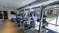 This fitness center is designed to provide a comprehensive workout environment within a single-level layout. The space is outfitted with a robust selection of exercise equipment, including multiple treadmills, elliptical cross-trainers with integrated digital displays, and a power rack loaded with free weights and barbells — catering to both cardio and strength training needs. A wall-mounted flat-screen television is positioned for easy viewing during workouts, adding an entertainment element to exercise sessions. Large windows with roller shades line one side of the room, allowing generous natural light to fill the space while offering views of an adjacent outdoor area, creating an open and energizing atmosphere. The flooring appears to be a durable, low-pile carpet suited for a fitness environment, and recessed lighting supplements the natural light for consistent illumination throughout. The layout is thoughtfully arranged to maximize usable floor space while maintaining clear pathways between equipment. The overall ambiance is clean, modern, and functional, making this fitness center a well-appointed facility for a variety of exercise routines.