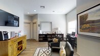 This single-floor space is designed with both comfort and functionality in mind. The main living area serves as a multifunctional common room, thoughtfully arranged to accommodate relaxation, reading, and productivity. The room features wood-look flooring throughout, which adds warmth and visual continuity to the space.

Seating is arranged around a patterned geometric area rug, with cushioned armchairs accented by decorative throw pillows, creating an inviting spot to unwind. A low, dark wood media console lines one wall, housing an extensive collection of books and media, making it a central feature of the room.

Adjacent to the seating area, a dedicated desk and computer workstation sits near a window, offering a bright, practical workspace. Natural light filters in through multiple windows dressed with white horizontal blinds, keeping the atmosphere light and airy.

The walls are finished in a clean, soft grey, complementing the neutral palette of the furnishings. Recessed lighting in the white textured ceiling provides even, ambient illumination throughout. A warm yellow-toned accent cabinet adds a touch of character near the entryway. Closet doors along the back wall suggest convenient built-in storage. The overall ambiance is calm, organized, and welcoming.