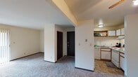 This single-floor unit offers a straightforward, functional layout designed for comfortable everyday living. The open living area is finished with wall-to-wall carpeting in a neutral gray tone, providing a soft, cohesive foundation throughout. A structural soffit detail above the entryway zone adds visual definition, helping to distinguish the living space from the entry corridor. Dark-stained, six-panel interior doors are featured consistently throughout, offering a grounding contrast against the light cream-colored walls and white baseboards.

The kitchen is efficiently arranged in a U-shaped configuration, maximizing counter and storage space. White cabinetry with warm wood-trimmed edges lines the walls, complemented by open display shelving above. The kitchen is equipped with a refrigerator, dishwasher, electric range, and a single-basin sink, all finished in white. Tile flooring in a stone-look pattern extends through the kitchen and into the bathroom, providing durability in high-traffic areas.

The two bedrooms are carpeted and bathed in natural light from windows fitted with horizontal blinds. Each room offers ample wall space for flexible furniture arrangement. The bathroom features matching tile flooring, a vanity with countertop, and a bi-fold dark-stained door that echoes the design language found throughout the unit.