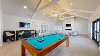 This single-floor community clubhouse in Clarksville, Indiana, is thoughtfully designed to offer a refined and inviting gathering space. The main lounge area commands attention with soaring vaulted ceilings accented by exposed white beams and elegant gold chandelier lighting. A sleek linear fireplace is mounted beneath a wall-mounted television, creating a warm focal point surrounded by light blue upholstered seating and contemporary side tables. Large French doors open to an outdoor area, flooding the space with natural light and providing easy indoor-outdoor access.

The adjoining billiards area continues the vaulted ceiling design and features a full-size pool table with a vibrant teal felt surface, complemented by additional wall-mounted screens and a bar-height seating nook with green upholstered stools. The space flows seamlessly from recreation to relaxation.

A separate kitchen and bar area rounds out the clubhouse, equipped with stainless steel appliances, white shaker-style cabinetry, and a generous white countertop island. Stylish burnt-orange leather bar stools with gold accents line the island beneath pendant lighting, making it ideal for casual dining or social gatherings. Light wood-tone tile flooring ties all areas together, contributing to the space's clean, modern aesthetic throughout.