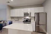 Located in the vibrant Georgetown neighborhood of Washington, DC, this well-appointed one-bedroom, one-bathroom residence offers a thoughtfully designed layout across a single floor. The modern kitchen is a standout feature, showcasing sleek, high-gloss white cabinetry extending to the ceiling, a suite of stainless steel appliances including a range, microwave, dishwasher, and refrigerator, and a clean white quartz island that doubles as additional prep space. Light wood-tone flooring flows seamlessly from the kitchen into the combined living and dining area, which is bathed in natural light from a large sliding glass door that opens to an outdoor view. The living and dining space offers a flexible, open layout well-suited for everyday living. The bedroom features plush carpeting, a walk-in closet, and its own sliding glass door providing additional natural light and outdoor access. The bathroom is finished with large-format light tiles, a floating vanity, and a glass-enclosed walk-in shower. Additional conveniences include a dedicated laundry area and a second closet for ample storage. The overall palette of soft grays, whites, and neutral tones creates a clean, contemporary ambiance throughout.