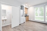 This single-floor unit offers a clean, modern design with warm wood-style laminate flooring and neutral gray tones throughout, creating a cohesive and functional living environment. The open main area serves as both a living and sleeping space, bathed in natural light from a large double window fitted with white horizontal blinds that frame views of surrounding greenery. Recessed lighting adds to the bright, welcoming ambiance.

The compact kitchenette is efficiently designed with light wood cabinetry, a dark countertop, an undermount sink, a built-in microwave, and a compact refrigerator — providing all essential amenities within a thoughtfully organized footprint. A tucked-away in-unit laundry closet houses a full stacked washer and dryer, adding a practical convenience rarely found in similarly sized spaces.

The entryway features a solid dark front door alongside an interior door providing access to the bathroom, with a closet nearby for storage. The bathroom is straightforward and functional, equipped with a combination tub and shower unit, a vanity sink, toilet, and a mirrored medicine cabinet.

Dark wood baseboards provide a subtle contrast to the light walls and flooring, lending the space a polished, finished look. Overall, this unit is well-suited for individuals seeking a self-contained, low-maintenance living space with modern finishes.