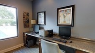 Located at 8000 Waters Ave in Savannah, Georgia, this single-floor business center offers a well-appointed and functional workspace designed to meet professional needs. The room features a long, granite-topped built-in desk that spans the width of the space, providing ample room for multiple workstations equipped with desktop computers, keyboards, and a shared printer — ideal for guests or tenants requiring office amenities. The cabinetry beneath the desk is finished in a rich dark wood tone, lending a polished, professional aesthetic to the room. The walls are painted in a cool, neutral gray that creates a calm and focused atmosphere, complemented by warm accent lighting from a classic table lamp. Decorative framed artwork adorns the walls, adding a creative and welcoming touch to the otherwise task-oriented environment. The flooring features a low-pile carpet that contributes to a quiet, distraction-free setting. Overall, the space strikes a balance between functional office utility and thoughtful interior design, making it a practical and comfortable environment for productivity.