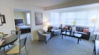 This well-appointed single-floor residence offers a warm and comfortable living environment designed with both function and style in mind. The open-plan living area is bathed in natural light, courtesy of a generous bay-style window arrangement fitted with white horizontal blinds, creating a bright and airy atmosphere throughout the day. Soft, neutral-toned wall-to-wall carpeting adds a sense of warmth and comfort underfoot, complementing the understated color palette of cream and light grey tones used throughout the space. The living area is thoughtfully arranged to accommodate both relaxed seating and a central coffee table, making it well-suited for everyday living. A coordinating accent chair and sofa provide comfortable seating, while decorative touches such as candlesticks, framed artwork, and potted greenery lend the space a refined, homely character. The bedroom is openly connected to the living area through a wide entryway, offering a seamless flow between spaces. The bedroom benefits from its own natural light source via an additional window, and features a substantial dark wood bed frame that anchors the room with a sense of elegance. Overall, the space presents a clean, inviting ambiance ideal for comfortable residential living.