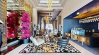 This expansive single-floor amenity space makes an immediate impression with its eclectic, high-design aesthetic. The main lounge area showcases soaring coffered ceilings, large-scale art installations, and an eye-catching geometric-patterned floor. Striking sculptural pieces — including a towering pink heart column — anchor the space alongside a mix of plush seating in bold stripes, leather, and velvet, all accented by gold sputnik chandeliers and globe pendant lights. A stylish reception and consultation area features a richly textured agate stone counter, deep navy blue walls, warm wood seating, and illuminated display cases. Intimate nooks throughout are finished with ornate damask wallcovering, marble-topped gold console tables, herringbone tile floors, and floor-to-ceiling window treatments that allow abundant natural light. A dedicated wet bar area incorporates mosaic stone tile backsplashes and mixed-material flooring, adding further textural depth. The state-of-the-art fitness center occupies a generously sized area with coffered ceilings, oversized windows with outdoor views, globe pendant lighting, and a full complement of modern cardio and strength equipment set against large-format athletic photography murals. Every zone of this space reflects a cohesive yet vibrant design language that balances luxury with energy.
