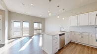 This thoughtfully designed one-bedroom, one-bathroom residence offers a modern aesthetic with quality finishes throughout. The open-concept kitchen and living area serves as the heart of the home, featuring a central island with a quartz countertop, white shaker-style cabinetry, and a full suite of stainless steel appliances including a refrigerator, range, microwave, and dishwasher. Pendant lighting above the island adds a stylish focal point, while light wood-tone flooring runs throughout the main living space, enhancing the bright, airy atmosphere. Large sliding glass doors lead to a private balcony, flooding the living area with abundant natural light and providing pleasant outdoor views. The bedroom is a comfortable retreat, finished with plush carpeting and a generously sized window that allows natural light to pour in. A dedicated closet offers practical storage. The full bathroom is clean and well-appointed, featuring white cabinetry with a dark countertop vanity, an undermount sink, a large mirror, and elegant pendant lighting. A hallway connects the private spaces with ease. Neutral warm-gray tones are used consistently throughout, lending the residence a cohesive and contemporary feel.