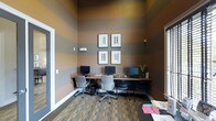 This single-floor business center is a well-appointed shared workspace designed to support productivity and convenience. The room's most striking design element is its bold, horizontally striped walls in alternating tones of warm terracotta and cool gray, which extend upward to notably tall ceilings, lending the space a sense of height and visual interest beyond its footprint.

A continuous built-in wooden desk runs along the back and side walls, accommodating three computer workstations, each equipped with a monitor, keyboard, and ergonomic task chair. The layout encourages efficient use of space while maintaining a degree of personal workspace for each user. A small decorative plant adds a touch of greenery near one end of the desk.

Four matching framed prints are arranged in a grid formation on the back wall, contributing a clean, professional aesthetic. Carpeted flooring in a muted, multi-toned pattern complements the color scheme while providing acoustic comfort. Natural light filters in through a window on the right side of the room, supplemented by wall-mounted lighting fixtures. A small reading or reference table is partially visible at the front of the space, offering additional utility for users.