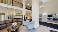 Located at 500 E Bruceton Rd in Pleasant Hills, PA, this well-appointed facility spans a generous single floor and offers a sophisticated, welcoming environment designed for comfort and community. The centerpiece of the space is a dramatic two-story lobby, characterized by soaring ceilings, prominent white columns, and a sweeping staircase with rich dark wood and wrought iron railings that overlook the main floor below. A tasteful chandelier adds an elegant focal point overhead, while the open-concept layout encourages a sense of airiness and flow throughout. The main seating area features a curated arrangement of upholstered sofas and armchairs around a classic wood and glass coffee table, creating an inviting gathering space. Adjacent to the lobby, a formal lounge area boasts floor-to-ceiling windows that flood the room with natural light, a built-in white fireplace with decorative mantle, and refined furnishings that contribute to a warm, residential ambiance. The dining area is bright and functional, fitted with granite-topped round tables, modern pendant lighting, and large windows offering outdoor views. A fully equipped kitchen includes stainless steel appliances, granite countertops, white cabinetry, and a central island, rounding out this thoughtfully designed communal facility.