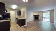 This single-floor residence offers a functional and cohesive layout with a clean, neutral palette throughout. The open-concept living and dining area is finished with light wood-tone laminate flooring and features a striking corner fireplace with a stone tile surround, serving as a natural focal point. A sliding glass door fills the space with natural light and provides direct access to a private balcony overlooking lush greenery. The kitchen is outfitted with dark espresso cabinetry, light countertops, stainless steel appliances including a range and refrigerator, and a pass-through window that connects to the dining area. A ceiling fan overhead adds to the functional design of this space. The bedrooms are carpeted for comfort, each featuring windows with blinds that bring in natural light and views of surrounding trees. One bedroom includes a spacious walk-in closet with built-in shelving. Baseboard heating is present in both bedrooms. The unit includes two bathrooms, each featuring dark vanity cabinetry with light countertops, a wall mirror, and wood-tone flooring. One bathroom offers a full soaking tub with a shower, while the other functions as a half-bath. Textured ceilings are consistent throughout the space, contributing to its cohesive residential character.