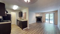 This single-floor residence offers a functional and cohesive layout with a clean, neutral palette throughout. The open-concept living and dining area is finished with light wood-tone laminate flooring and features a striking corner fireplace with a stone tile surround, serving as a natural focal point. A sliding glass door fills the space with natural light and provides direct access to a private balcony overlooking lush greenery. The kitchen is outfitted with dark espresso cabinetry, light countertops, stainless steel appliances including a range and refrigerator, and a pass-through window that connects to the dining area. A ceiling fan overhead adds to the functional design of this space. The bedrooms are carpeted for comfort, each featuring windows with blinds that bring in natural light and views of surrounding trees. One bedroom includes a spacious walk-in closet with built-in shelving. Baseboard heating is present in both bedrooms. The unit includes two bathrooms, each featuring dark vanity cabinetry with light countertops, a wall mirror, and wood-tone flooring. One bathroom offers a full soaking tub with a shower, while the other functions as a half-bath. Textured ceilings are consistent throughout the space, contributing to its cohesive residential character.