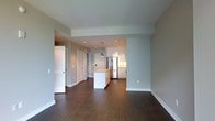 This single-floor space showcases a thoughtfully designed open-concept layout that seamlessly connects the living area to the kitchen. Dark hardwood floors run throughout, providing a rich contrast against the light grey walls and white trim that define the interior aesthetic.

The kitchen is a standout feature, equipped with white cabinetry, glossy upper cabinets that extend toward the ceiling, and stainless steel appliances including a full-size refrigerator. A peninsula with a white waterfall-style counter serves as both a workspace and casual dining area. A pendant light fixture hangs above, adding a decorative focal point. Recessed lighting illuminates the kitchen zone effectively.

The living area is generously proportioned, offering flexibility in furniture arrangement. The neutral wall tones and consistent flooring create a cohesive, airy feel throughout the space. A hallway visible from the main area leads to additional rooms, flanked by multiple doors suggesting dedicated storage or secondary spaces. White baseboards and clean architectural lines contribute to the overall modern, well-maintained character of the interior.