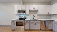 This single-floor unit at 2176 E 105th St in Cleveland, Ohio presents a thoughtfully designed layout that maximizes its compact footprint. Light wood-tone vinyl plank flooring flows consistently throughout the space, complemented by crisp white walls, recessed lighting, and dark hardware accents that provide visual contrast.

The kitchen is a standout feature, outfitted with two-tone cabinetry—white upper cabinets and gray lower cabinets—paired with white countertops and a full suite of stainless steel appliances, including a range, over-the-range microwave, dishwasher, and refrigerator. A single-basin sink with a matte black faucet adds a modern touch.

The open main living area is bright and airy, with a window allowing natural light to filter in. Recessed ceiling lights ensure even illumination throughout. A dedicated in-unit laundry closet with louvered doors houses a stacked washer and dryer, adding significant convenience.

The bathroom features light wood-tone flooring, a walk-in shower with a bold black-framed sliding glass door, a white vanity with light countertop, and matte black fixtures, creating a clean, contemporary feel. An additional flexible room provides space adaptable to various needs.