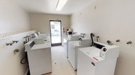 This single-floor shared laundry facility is designed for practical, high-capacity use. The room is arranged with rows of top-loading washing machines lining both sides, creating a clear central aisle for easy access and movement throughout the space. At the back of the room, stacked commercial-grade dryer units are positioned against the wall, maximizing the available floor space and accommodating multiple users simultaneously.

The interior features neutral beige and white walls that contribute to a clean, utilitarian aesthetic. Overhead fluorescent lighting provides bright, even illumination across the entire room. The floor is finished with a durable, light-toned vinyl surface suited for a high-traffic environment.

A glass-panel entry door is located along one wall, allowing natural light to filter into the space and providing a view of the surrounding outdoor area. Adjacent to the entry door, a wall-mounted fire extinguisher and a waste receptacle are conveniently positioned for safety and cleanliness.

Additional utility fixtures, including faucet connections along the walls, indicate thoughtful infrastructure planning throughout the space. A security camera is also visible, ensuring a monitored and secure environment for users. Overall, the facility is well-organized, efficiently laid out, and equipped to serve the laundry needs of a shared community or residential setting.