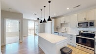 Located at 135 Ballentrae Ct in Chapel Hill, NC, this well-designed one-bedroom, one-bathroom residence offers a thoughtfully laid-out single-level floor plan. The heart of the home is a modern open-concept kitchen, showcasing crisp white shaker cabinetry, light quartz countertops, and a subway tile backsplash. A large center island with built-in shelving and trio of matte black pendant lights serves as both a functional workspace and a visual focal point. Stainless steel appliances, including a refrigerator with ice maker, electric range, microwave, and dishwasher, complete the kitchen. Warm hardwood-style flooring flows throughout the main living areas, while recessed lighting and a ceiling fan contribute to a bright, airy ambiance. A stacked in-unit washer and dryer adds everyday convenience. The bedroom offers plush carpeting, natural light through blinds-fitted windows, and access to a dedicated closet. The full bathroom features a tiled tub-shower combination with a built-in niche, hardwood-style flooring, and clean, neutral finishes. A private balcony accessible from the living area provides an outdoor retreat with open views of the surrounding area.