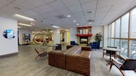 This single-floor space is thoughtfully designed to accommodate a variety of activities and social interactions across several distinct zones. The main lounge area serves as a central gathering point, anchored by a striking stone fireplace with a bold red mantel, flanked by vivid blue velvet armchairs and a golden ottoman, creating a warm and inviting focal point. Adjacent to the fireplace, a seating cluster features rich leather sofas and mid-century-style chairs arranged on wood-look tile flooring, ideal for casual conversation.

A standout feature of the open common area is a collection of freestanding wooden hanging chairs, adding a playful and distinctive design element. This zone flows into a collaborative area with a high-top conference table illuminated by an industrial-style chandelier, set atop patterned green and grey carpet tiles that define different functional areas throughout the space.

A bright lounge area with modular sofas and a dedicated computer workstation row offers both relaxation and productivity options. A reception-style counter with a bold yellow accent wall and pendant lighting adds energy to the entry zone. Additional flexible rooms with open shelving and modular seating provide adaptable spaces for various uses. Recessed lighting and a drop ceiling are consistent throughout, contributing to a clean, modern aesthetic.